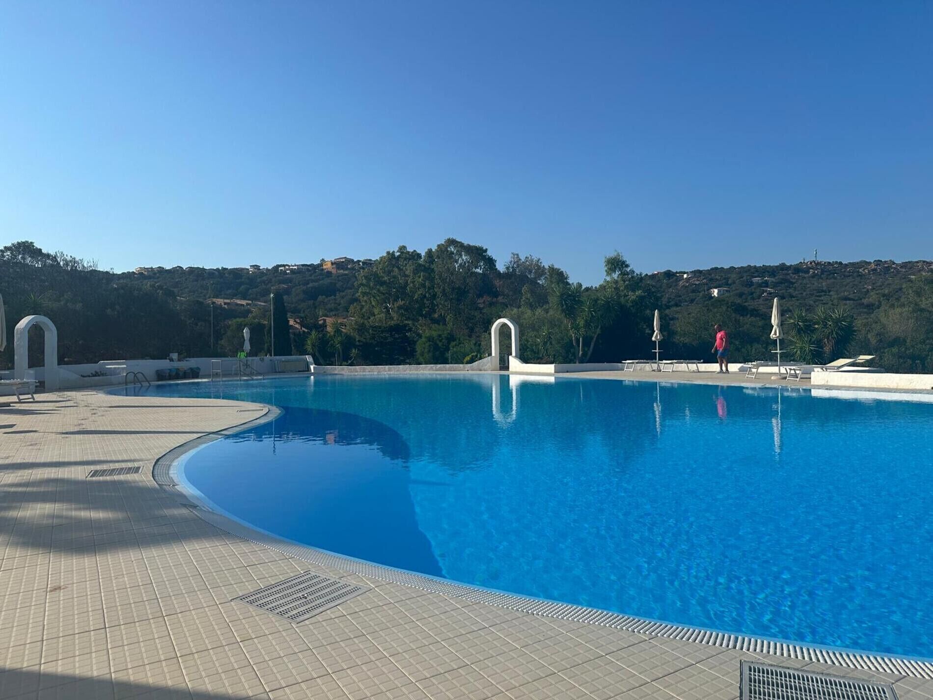 A clear blue swimming pool with a tiled deck, surrounded by greenery and hills in the background.