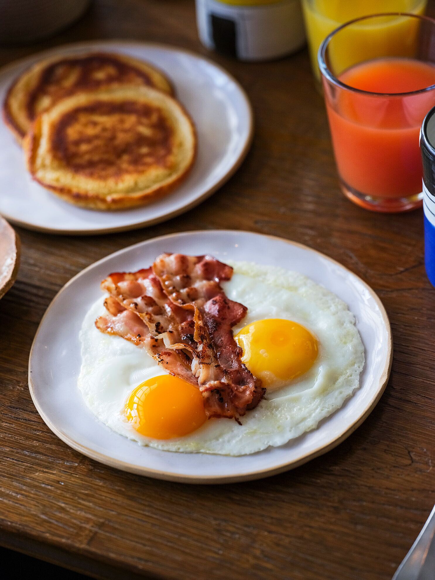 A breakfast plate with eggs and bacon, a glass of orange juice, and pancakes on a wooden table.