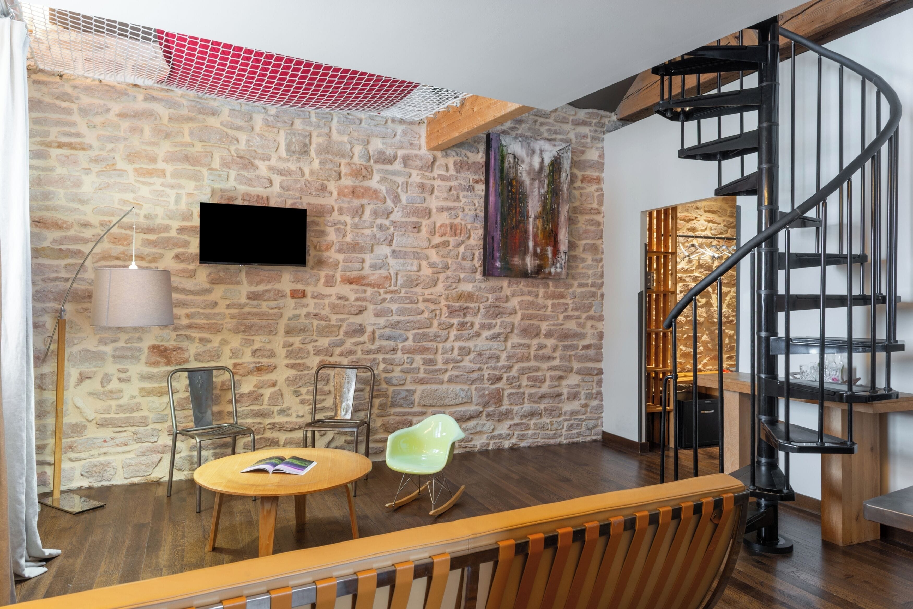 A room with a stone wall, a flat-screen TV, a wooden table, two chairs, a green chair, a spiral staircase, and a hanging light fixture.