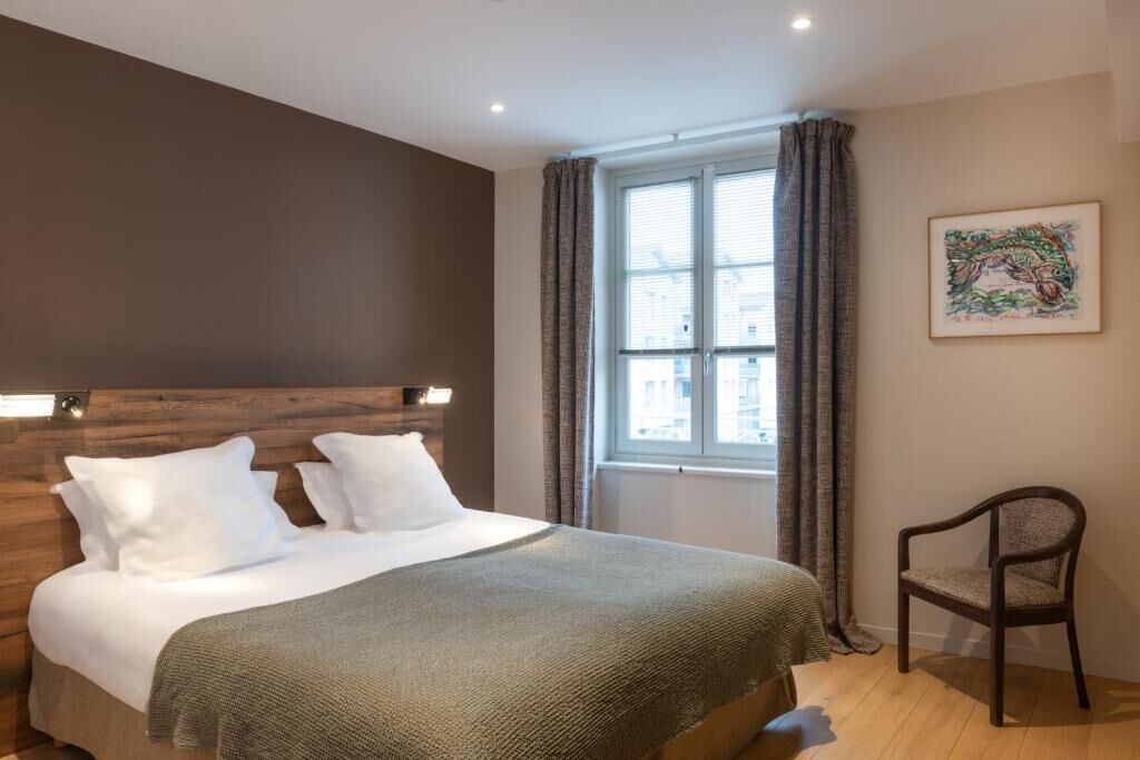 A hotel room with a bed, a window with curtains, a chair, and a wall-mounted artwork.