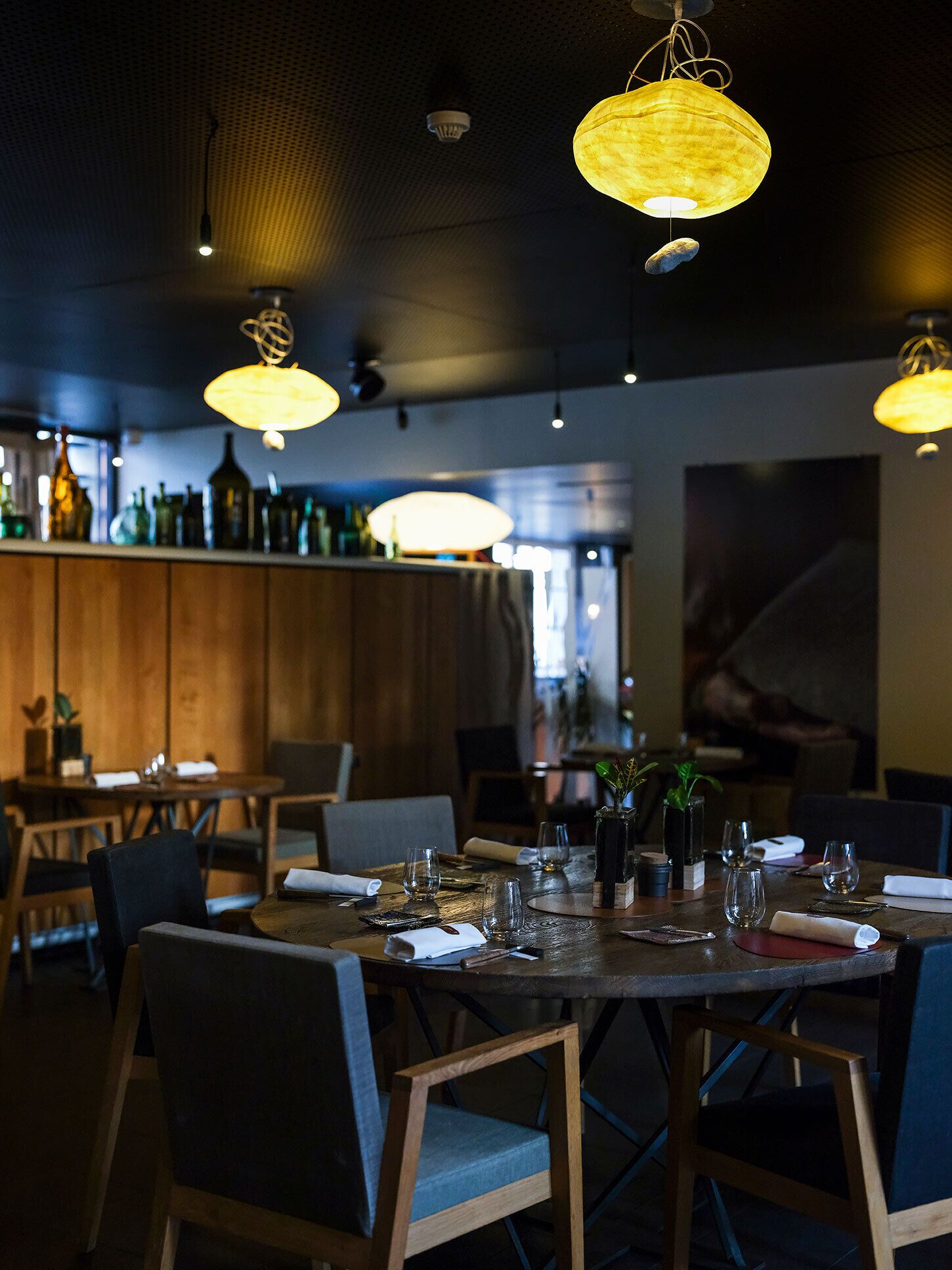 A restaurant interior with round tables, chairs, hanging lamps, and a bar area in the background.