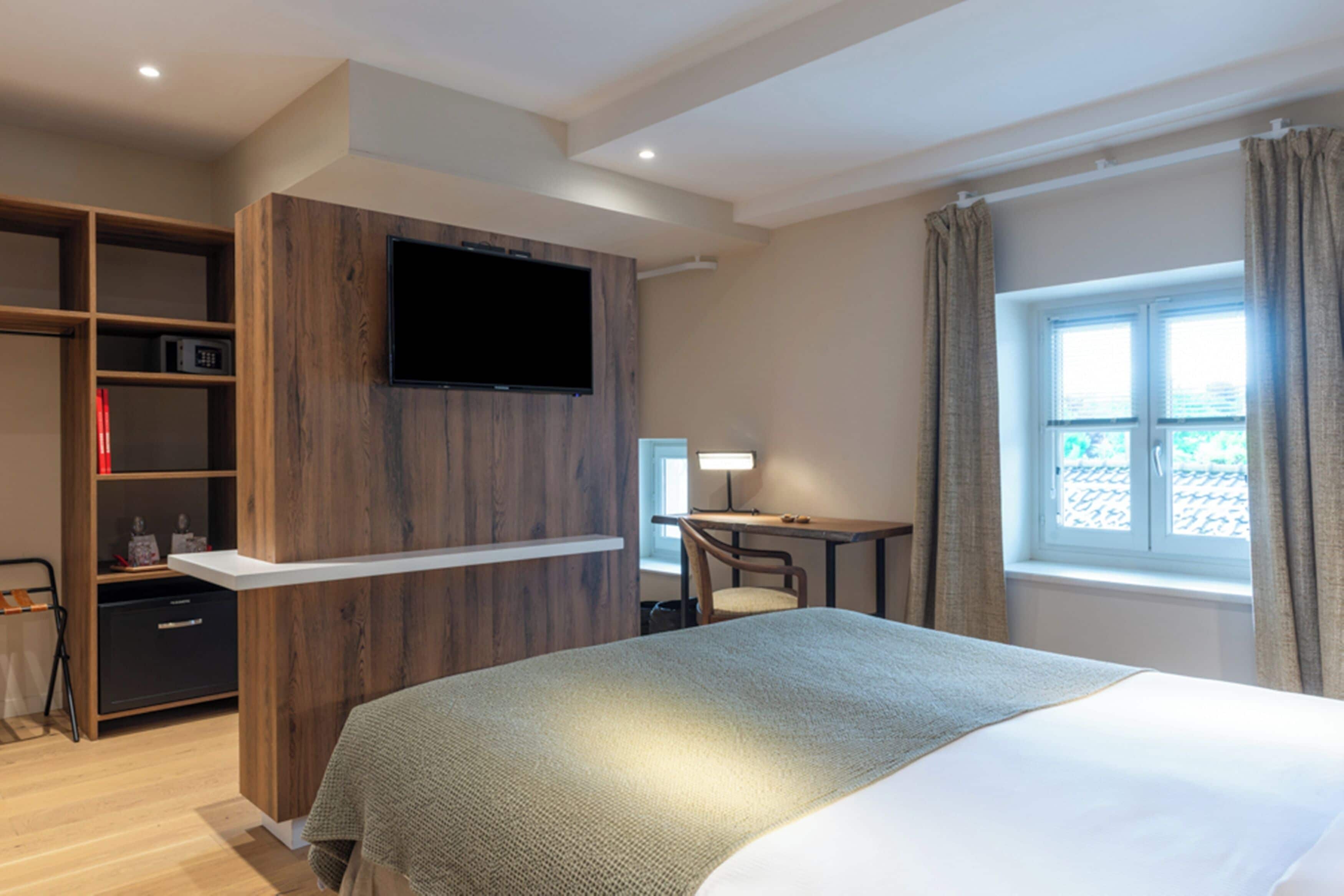 Quarto de hotel com cama, escrivaninha, televisão presa a um painel de madeira, guarda-roupa e janela com cortinas.