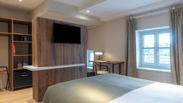 Quarto de hotel com cama, escrivaninha, televisão presa a um painel de madeira, guarda-roupa e janela com cortinas.