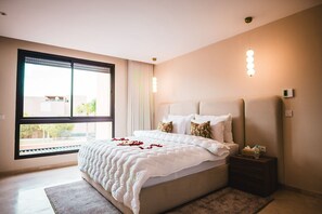 A modern bedroom with a large bed, a nightstand, a window with blinds, and a wall-mounted light fixture. - Riad azur Al Maaden Golf – Marrakech (Marrakech)