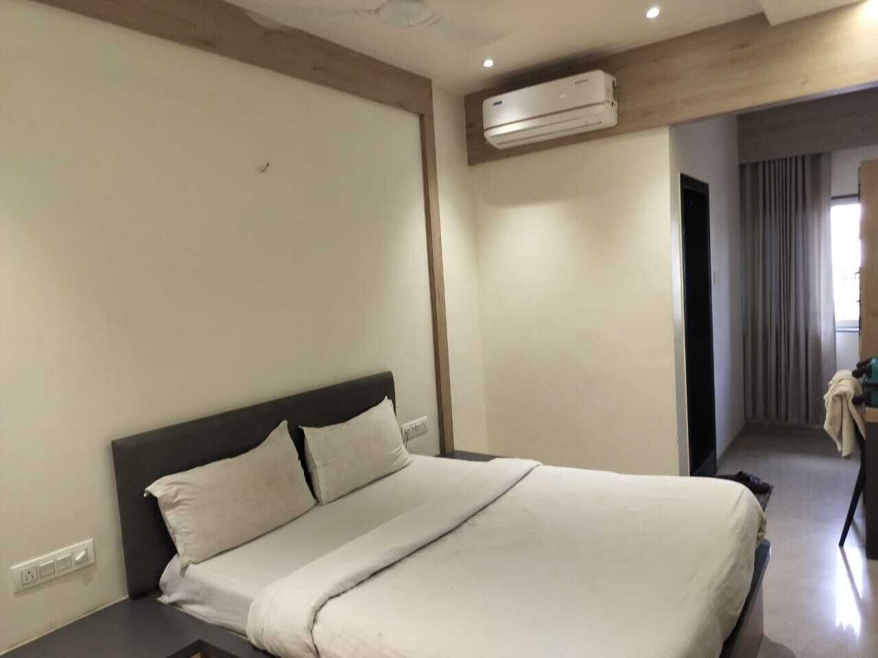 A hotel room with a bed, a bedside table, an air conditioning unit, and a view of a corridor with a door.