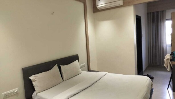 Habitación de hotel con cama, mesita de noche, equipo de aire acondicionado y vista a un pasillo con una puerta.
