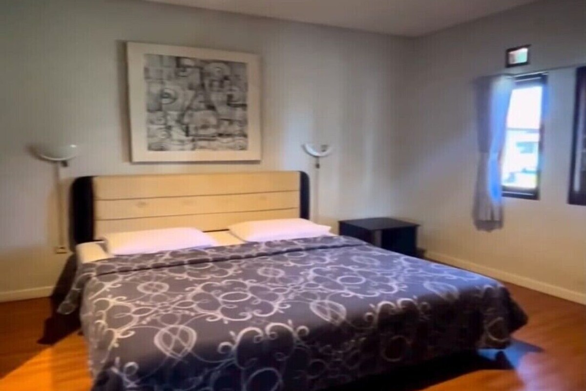 Ein Schlafzimmer mit einem Bett, einem Nachttisch, einer Wandlampe und einem Fenster mit Vorhängen.