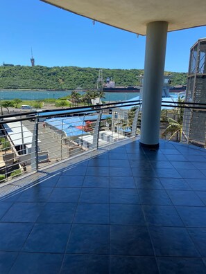 Un balcón con vista a un puerto, un barco grande y una torre a lo lejos.