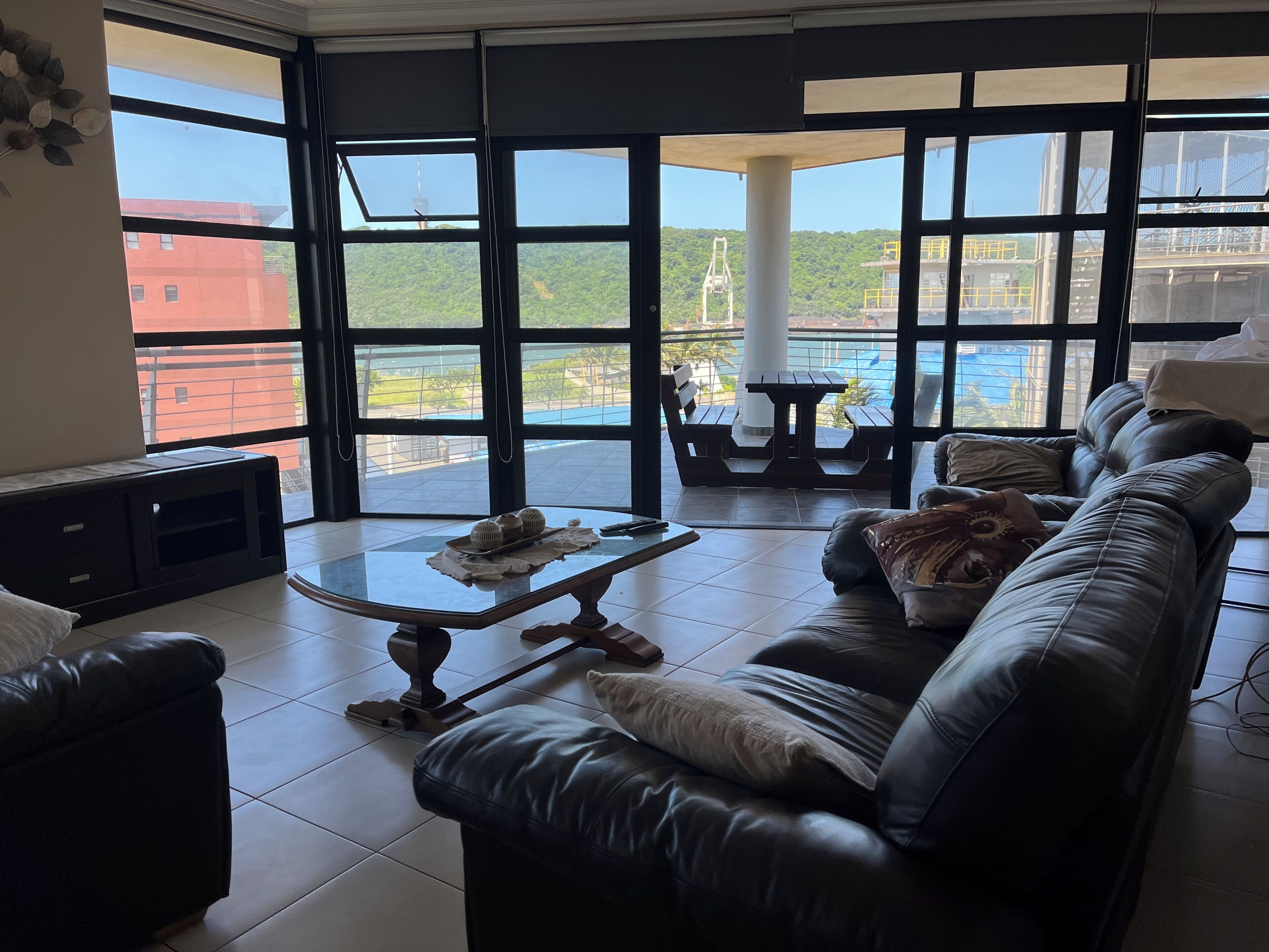A spacious living area with a leather sofa, a glass coffee table, and a view of a water body and greenery through large windows.