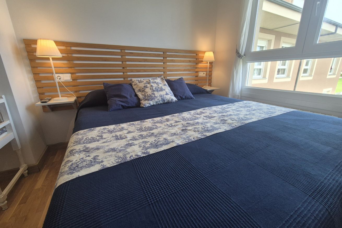 Ein Schlafzimmer mit einem Holzkopfteil, zwei Nachttischlampen, einem Bett mit blauen und weißen Kissen und einem Fenster mit Blick auf Gebäude.