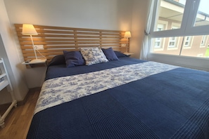 A bedroom with a wooden headboard, two bedside lamps, a bed with a blue and white patterned bedspread, and a window with a view of buildings. - 45A01 Apartment in Puerto de Vega (Puerto de Vega)
