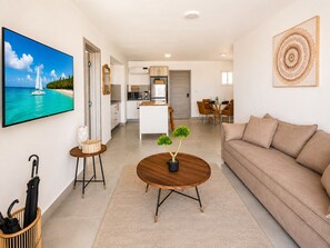 Ein stilvolles Wohnzimmer mit einer Couch, einem Couchtisch mit Zierpflanzen, einem Essbereich mit Tisch und Stühlen, einer Küche mit Schränken und Geräten sowie einem Fernseher, der eine schöne Aussicht zeigt.