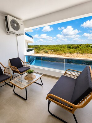 Ein Balkon mit Korbmöbeln, einem Glastisch mit Pflanze sowie Blick auf einen Pool und Grünflächen.