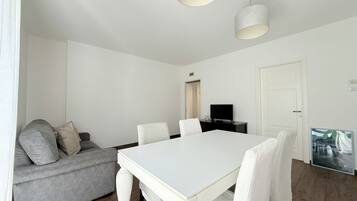 A modern living room with a white dining table, grey sofa, and a flat-screen TV.