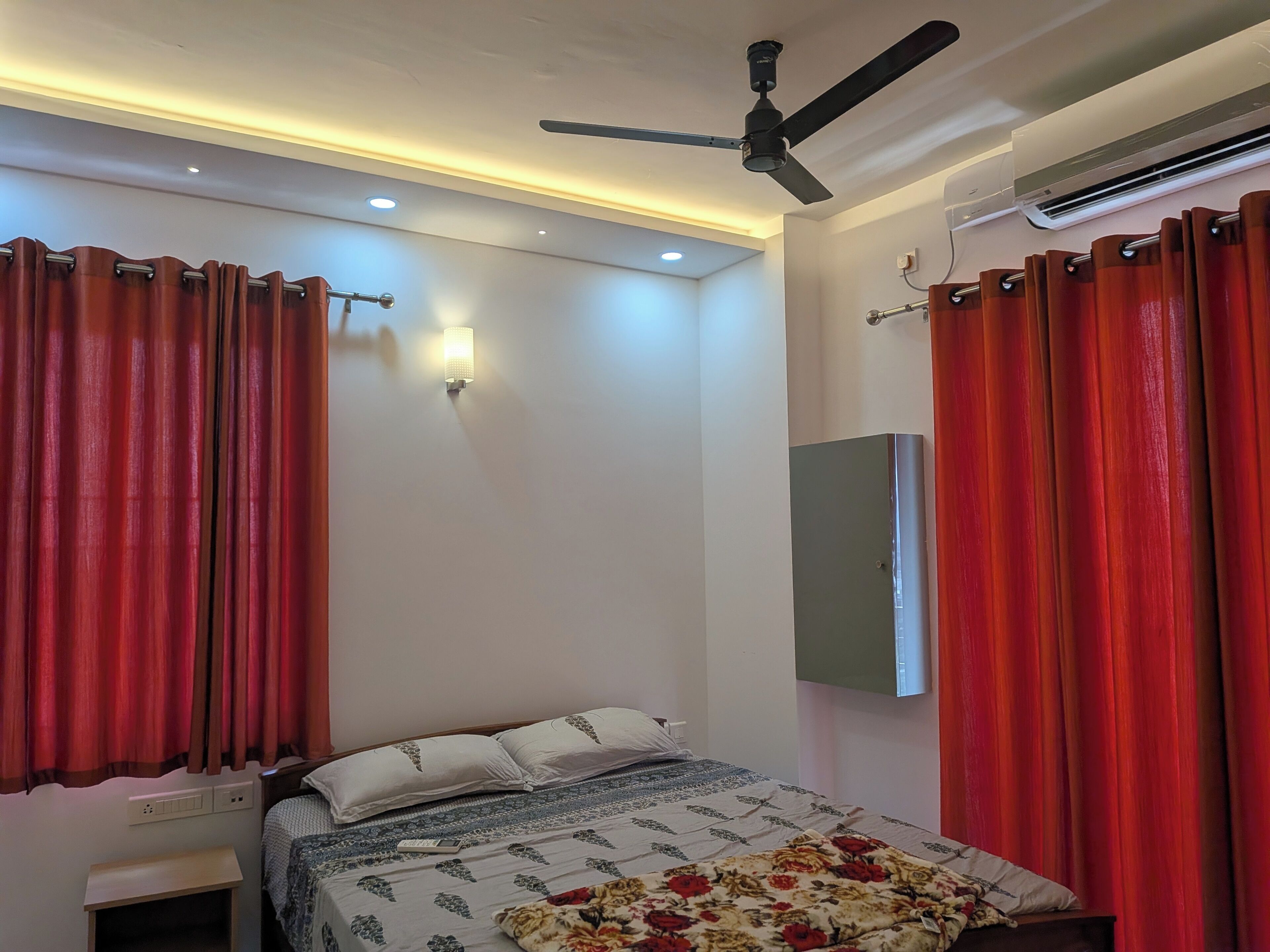 A bedroom with a bed, curtains, a ceiling fan, an air conditioning unit, and a wall-mounted light fixture.
