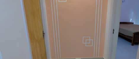 A hallway with a wooden door, a wall with geometric patterns, and a bed in the background.