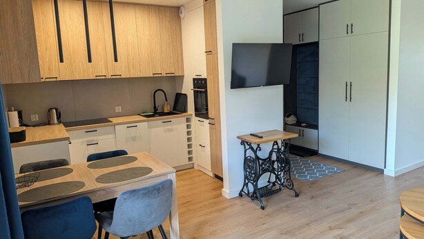 A modern kitchen with wooden cabinets, a dining area with a table and chairs, and a small side table. - Klonova Apartment in Masuria (Olecko)
