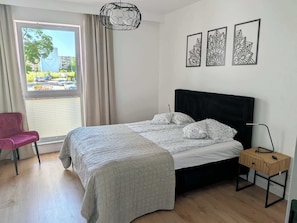 A bedroom with a bed, a chair, a bedside table, and a window with curtains.