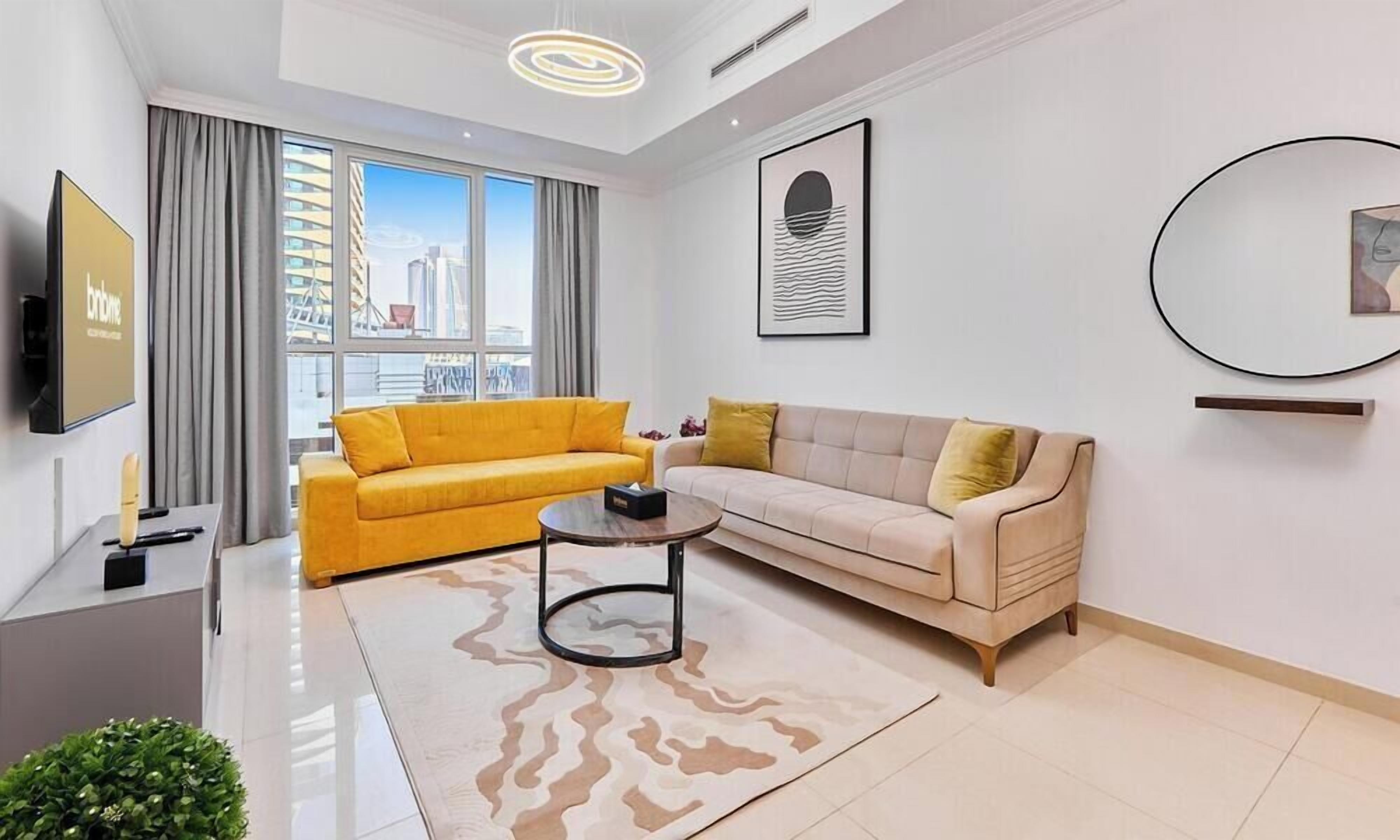 Un salon moderne avec un canapé jaune, un canapé beige, une table basse et une grande fenêtre donnant sur la ville.