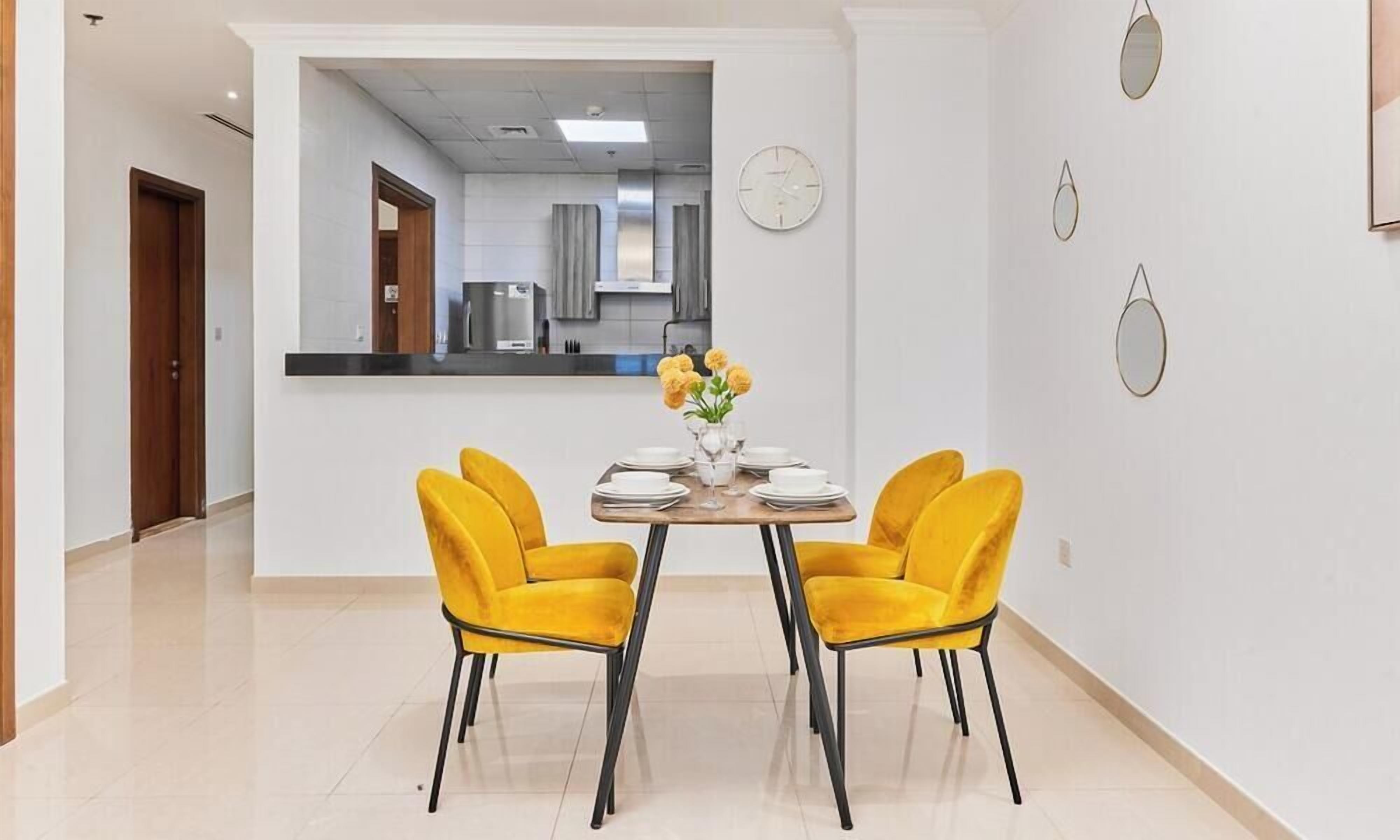 Comedor con una mesa lista para dos personas, sillas amarillas y un florero con flores.