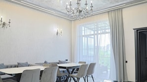 A dining room with a round table, grey upholstered chairs, a chandelier, and a large window with curtains. - Leader Hotel (Goris)