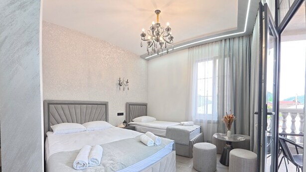 A hotel room with two beds, a chandelier, a small table with a vase, and a view of the outside. - Leader Hotel (Goris)