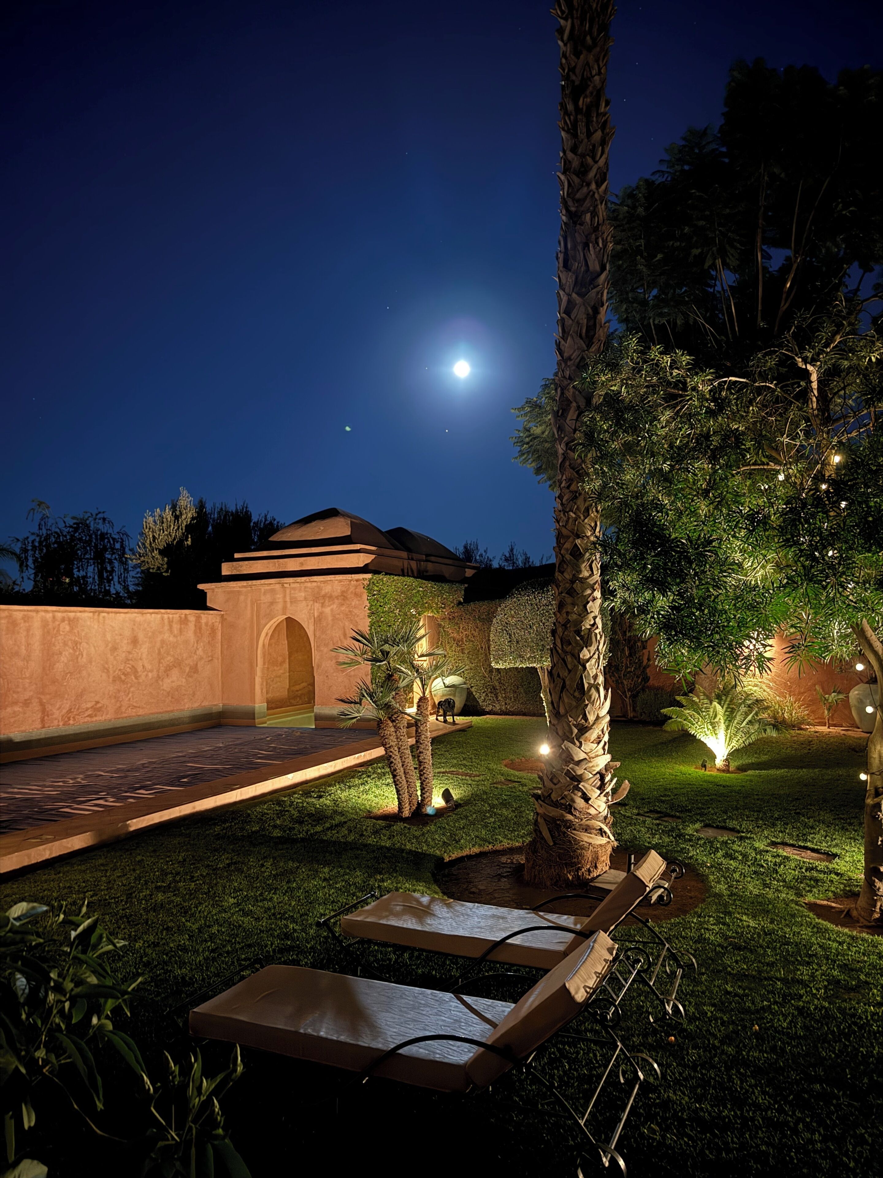 A nighttime outdoor area with a pool, lounge chairs, palm trees, and a building with an arched entrance.