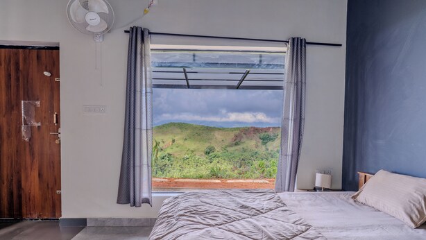 Un dormitorio con cama, ventana con cortinas, ventilador de pared y vista a un paisaje verde.