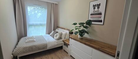 A bedroom with a bed, bedside table, dresser, window with curtains, and a wall-mounted picture.