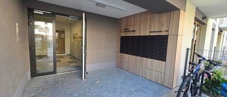 A modern apartment building entrance with a glass door, a wooden wall with apartment numbers, and a bicycle parked outside.