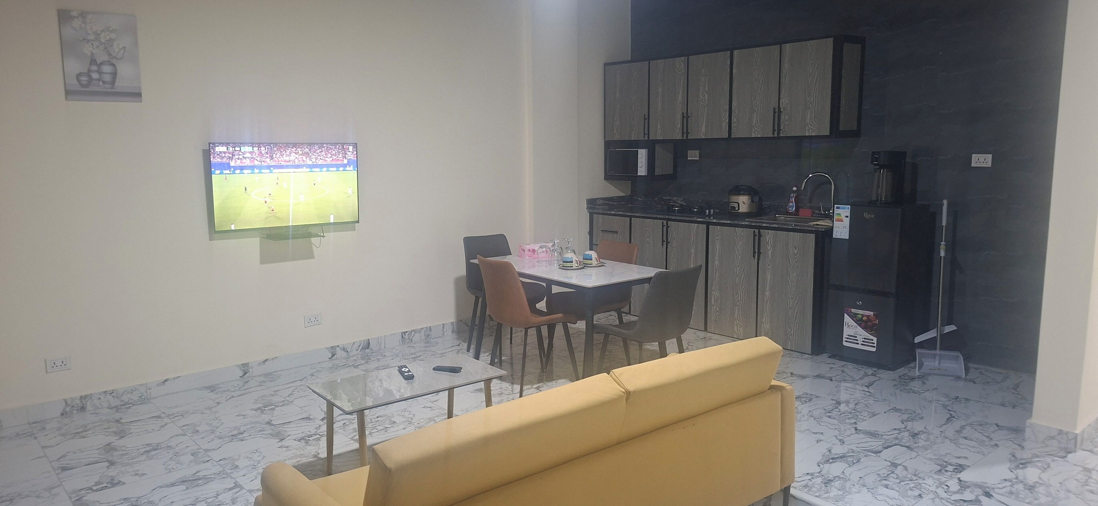 A compact living space with a dining area, a sofa, a kitchen with a microwave, and a TV displaying a sports event.