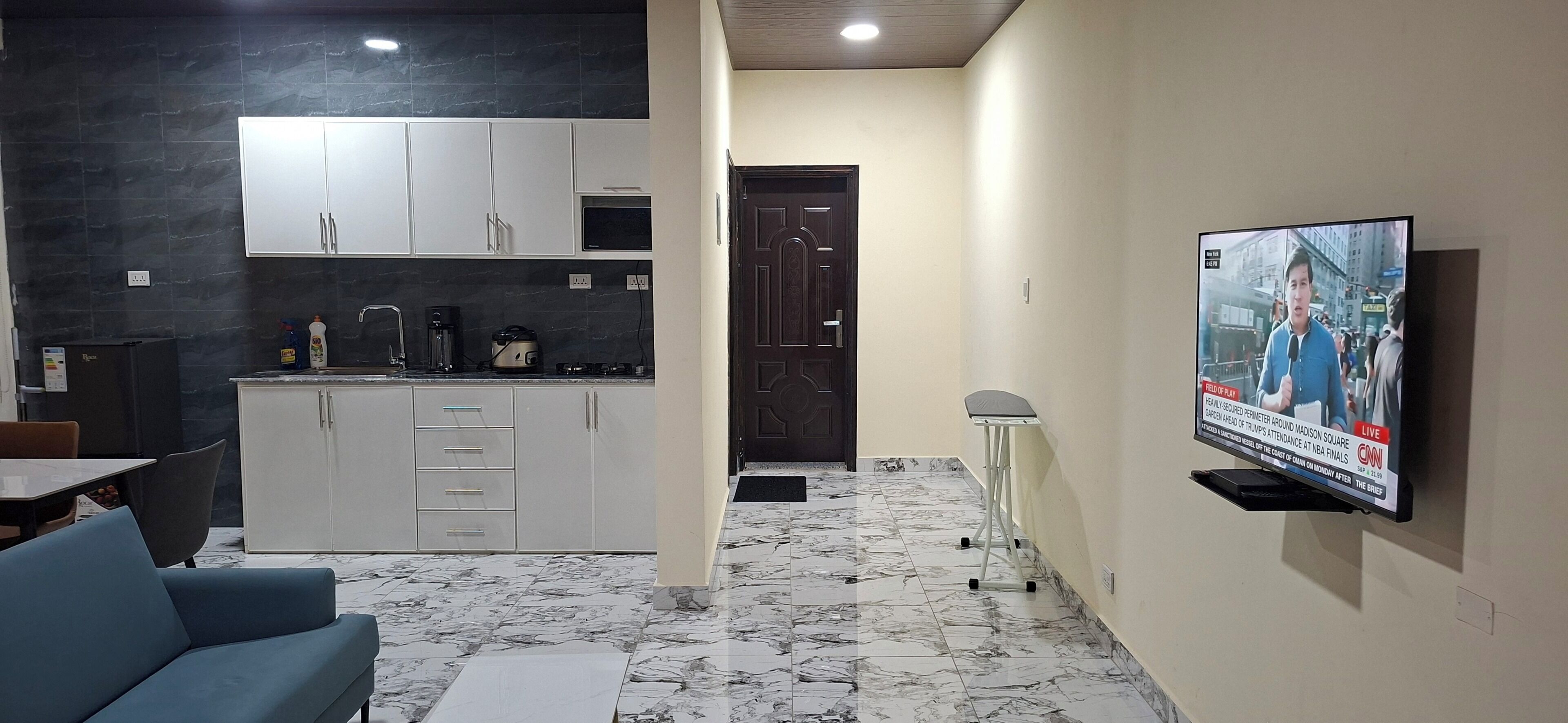 Una sala de estar moderna con un televisor mostrando un noticiero, una zona de cocina con armarios blancos y un suelo con estampado de mármol.