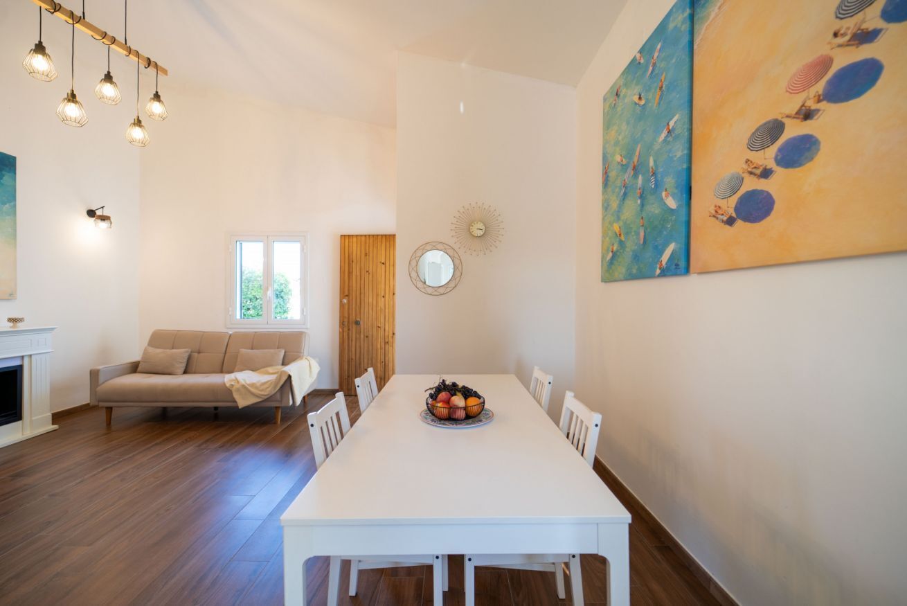 A dining area with a white table, chairs, and a fruit bowl centerpiece. There is a wooden door and a window with a view of greenery.