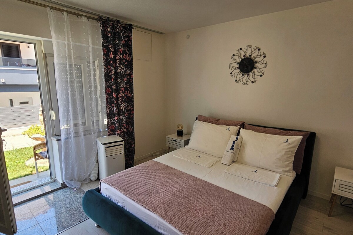 Ein Schlafzimmer mit Bett, Nachttisch, Wanddekoration, Fenster mit Vorhängen und Blick ins Freie.