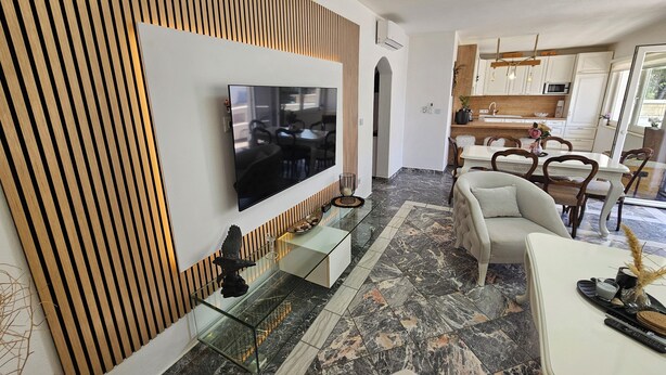 A modern living room with a flat-screen TV mounted on a wooden panel wall, a glass coffee table, and a dining area with a white table and chairs. - Casa Carmel (Kamen)