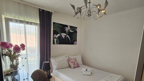 A bedroom with a single bed, a bedside table, a chair, a window with curtains, a painting, and a chandelier. - Casa Carmel (Kamen)