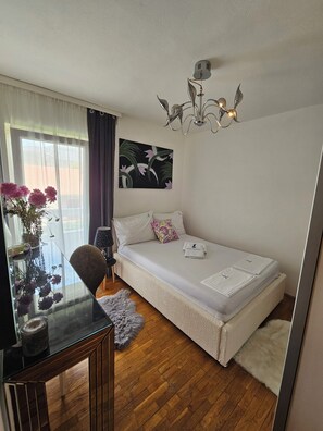 Ein Schlafzimmer mit einem Einzelbett, einem Nachttisch, einem Sessel, einem Fenster mit Vorhängen, einem Gemälde und einer Deckenleuchte.