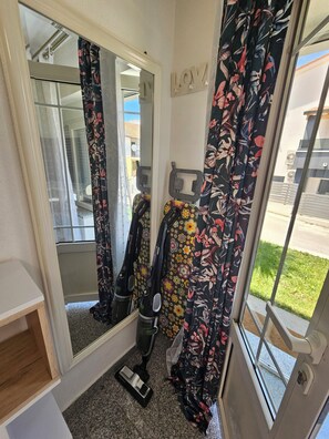 Ein Zimmer mit einem großen Spiegel, einem Staubsauger, blumigen Vorhängen und Blick ins Freie.