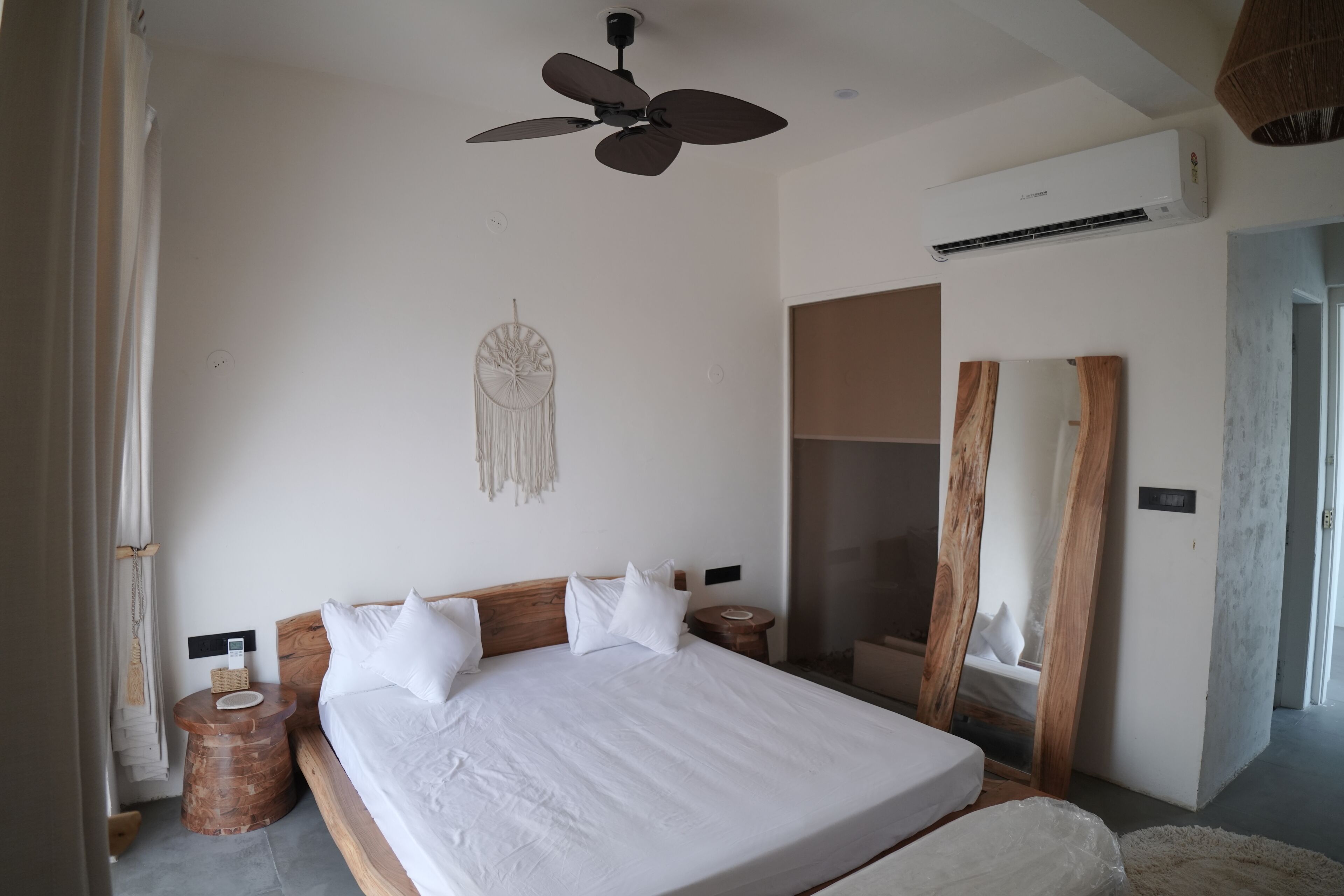 A bedroom with a large bed, wooden headboard, bedside tables, a ceiling fan, an air conditioning unit, and a wall-mounted decorative piece.