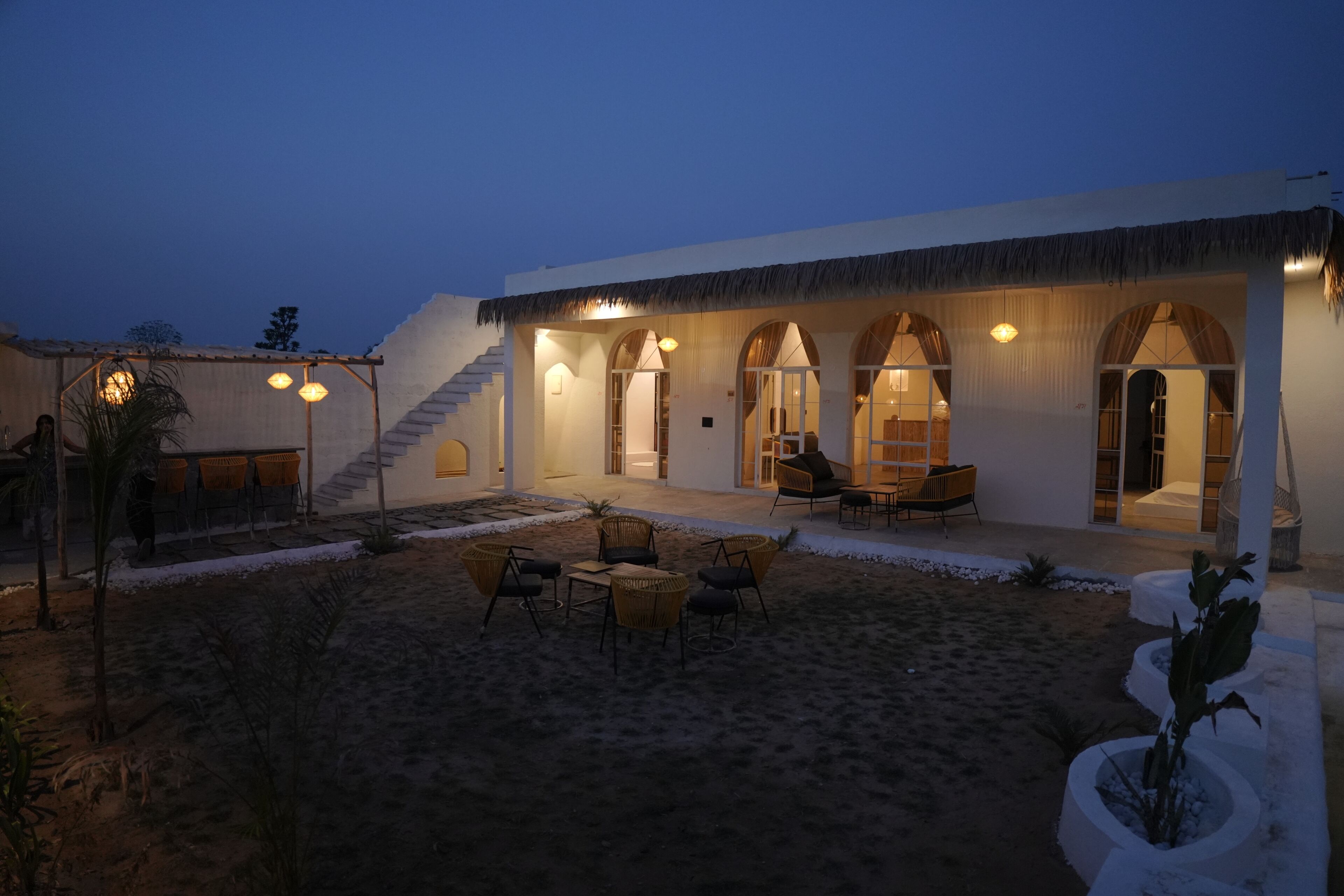 A building with a covered outdoor seating area, a staircase leading to an upper level, and a sandy courtyard.