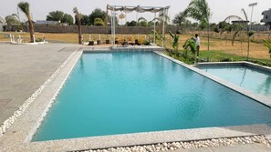 A rectangular swimming pool with a stone border, surrounded by a paved area with chairs and a covered seating area.