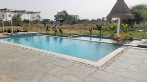 A rectangular swimming pool with a stone border, surrounded by a patio area with chairs and a thatched umbrella. - Mahika Valley Stay (Jaipur)