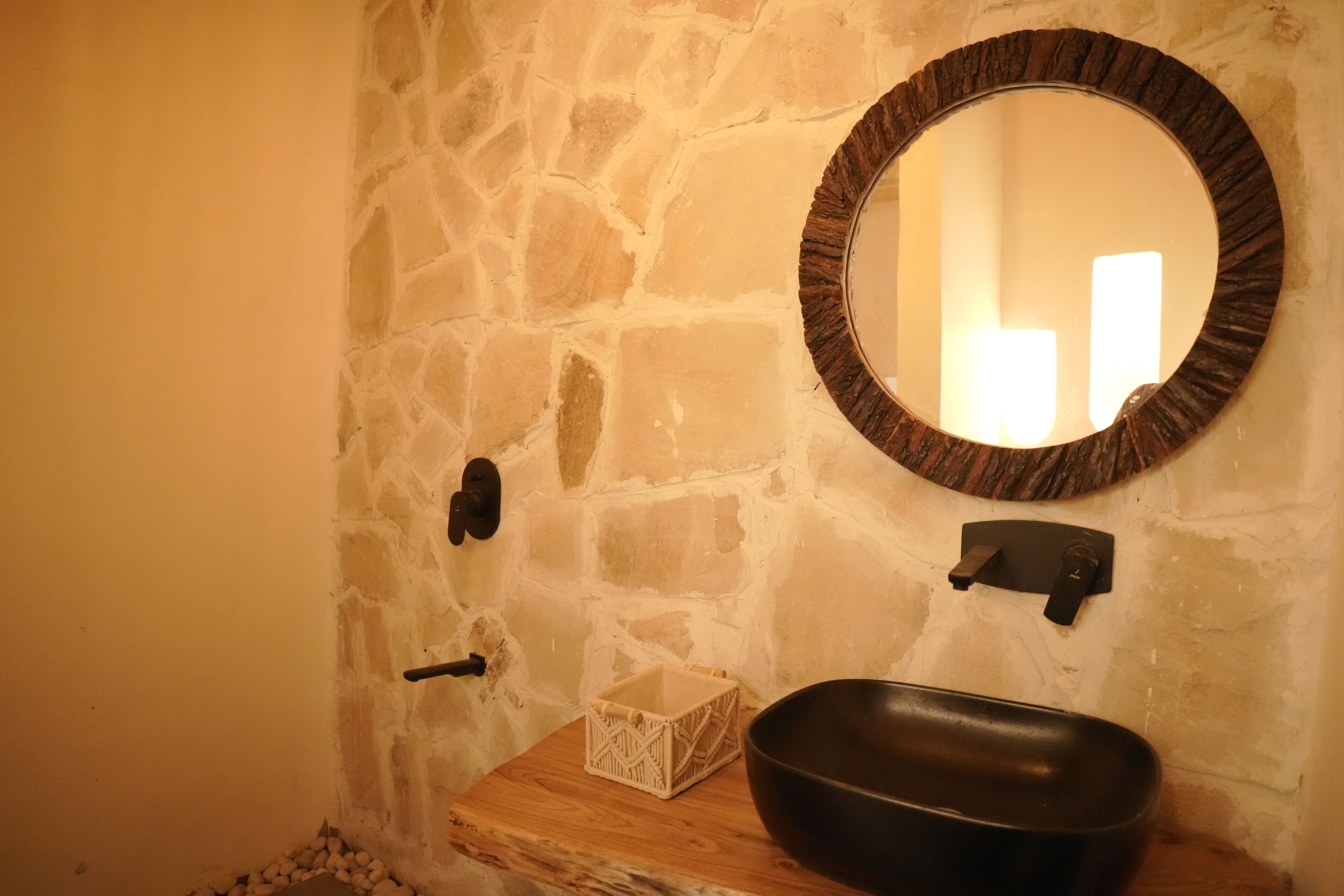Un baño con pared de piedra, un espejo redondo, un lavabo negro y una repisa de madera.
