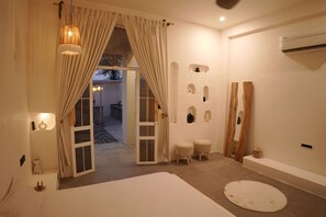 A room with a bed, a bathtub, and a small seating area. There are curtains, a wall-mounted air conditioner, and a door leading outside.