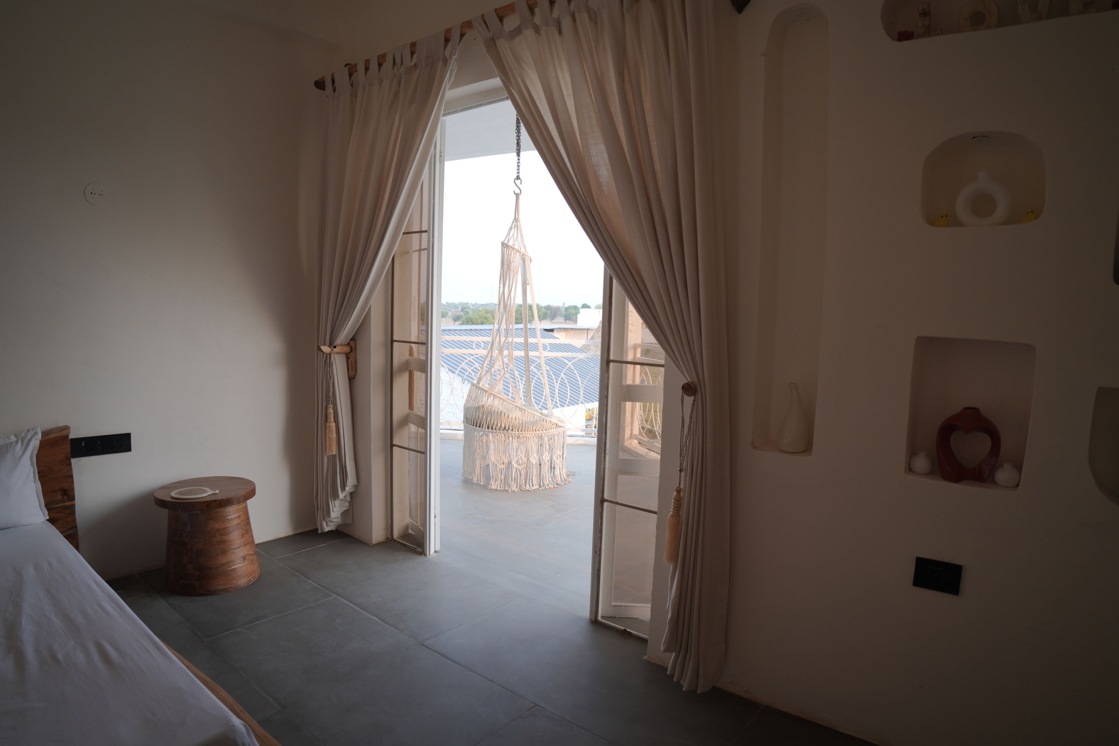 A room with a bed, a round wooden table, and a macrame chair. There are curtains on the windows, and a view of the outside.