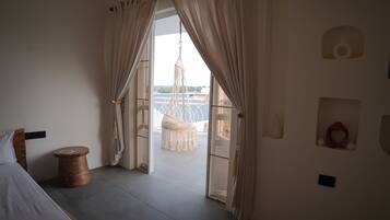 A room with a bed, a round wooden table, and a macrame chair. There are curtains on the windows, and a view of the outside.