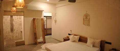 Un dormitorio con cama, ventilador de techo, cabecero de madera, atrapasueños colgado en la pared y pared de piedra.