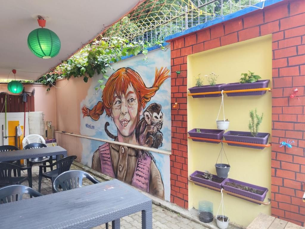 Un murale raffigura una persona e un cane, con sedute all'aperto che includono tavoli e sedie, e un muro in mattoni con mensole piene di piante in vaso.