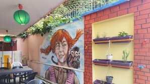 Um mural com uma pessoa e um cão, mesas e cadeiras ao ar livre, e uma parede de tijolos com prateleiras cheias de plantas em vasos.