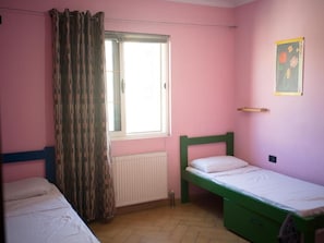 A room with two single beds, a window with curtains, a radiator, and a wall-mounted shelf.